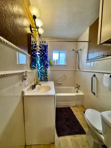 Wall-mounted vanity light fixture, built-in bathtub with shower, window, single vanity with integrated sink, and toilet with a white finish