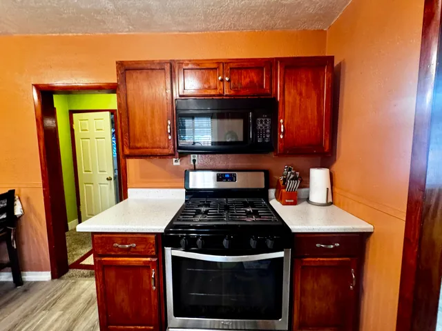 Wood-finish cabinetry with silver hardware, white speckled countertops, built-in microwave, and stainless steel gas range