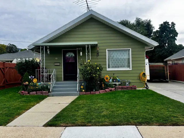 Gabled roofline with green siding, concrete front walkway, covered porch with decorative metal railing, and a side driveway