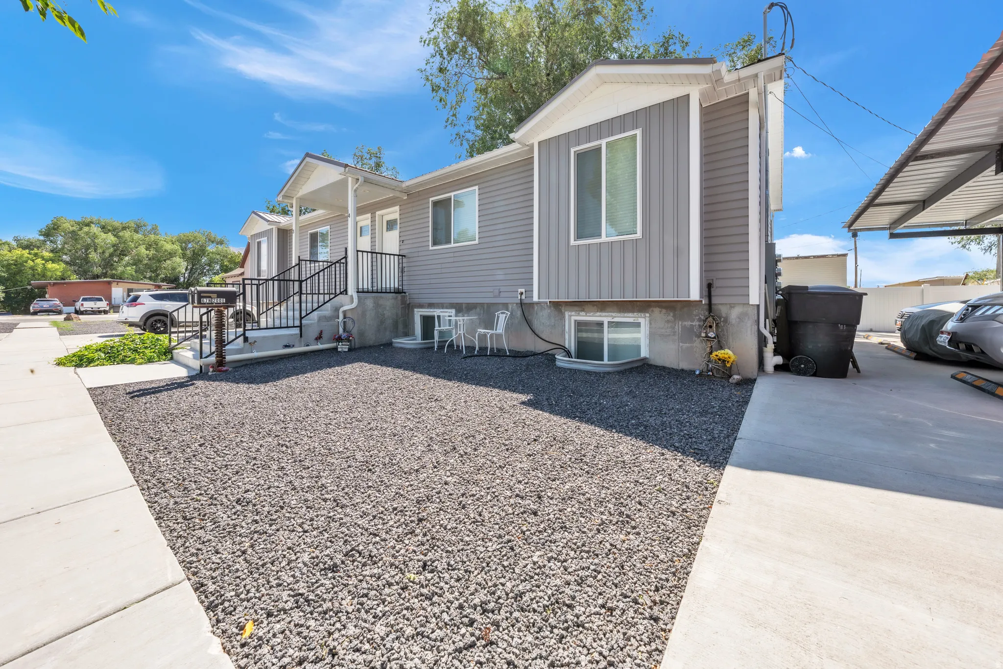 Gray siding exterior with white trim, front porch with black railing, concrete walkway, gravel landscaping, and a covered carport