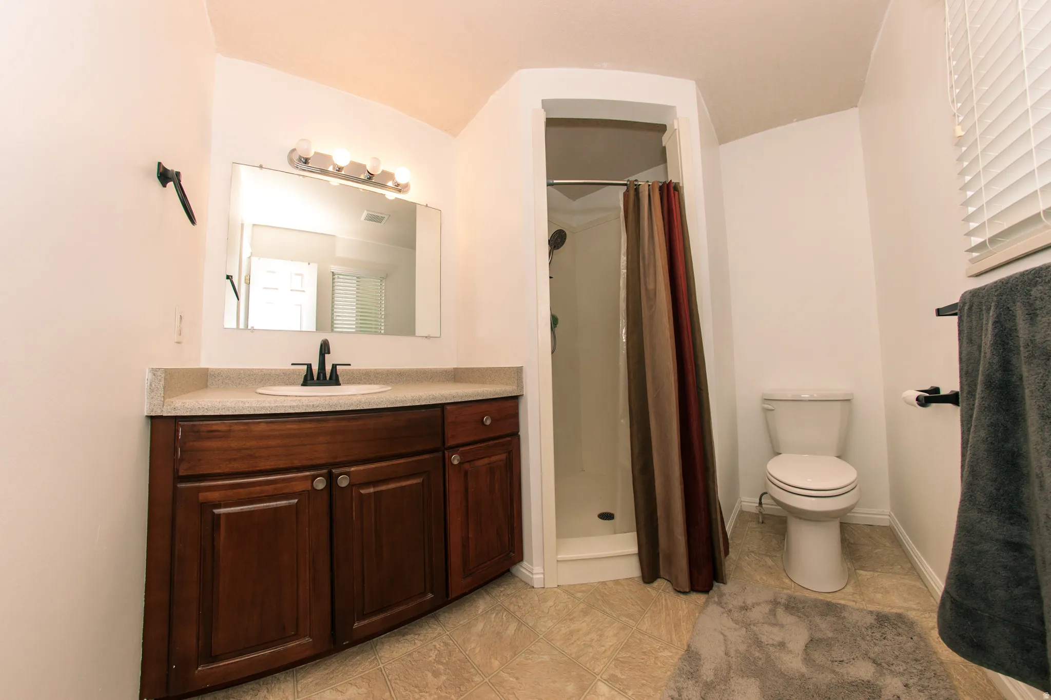 Bathroom featuring a wood-finish vanity with a light-toned countertop, an integrated sink, a rectangular mirror, and a standing shower with a curved enclosure