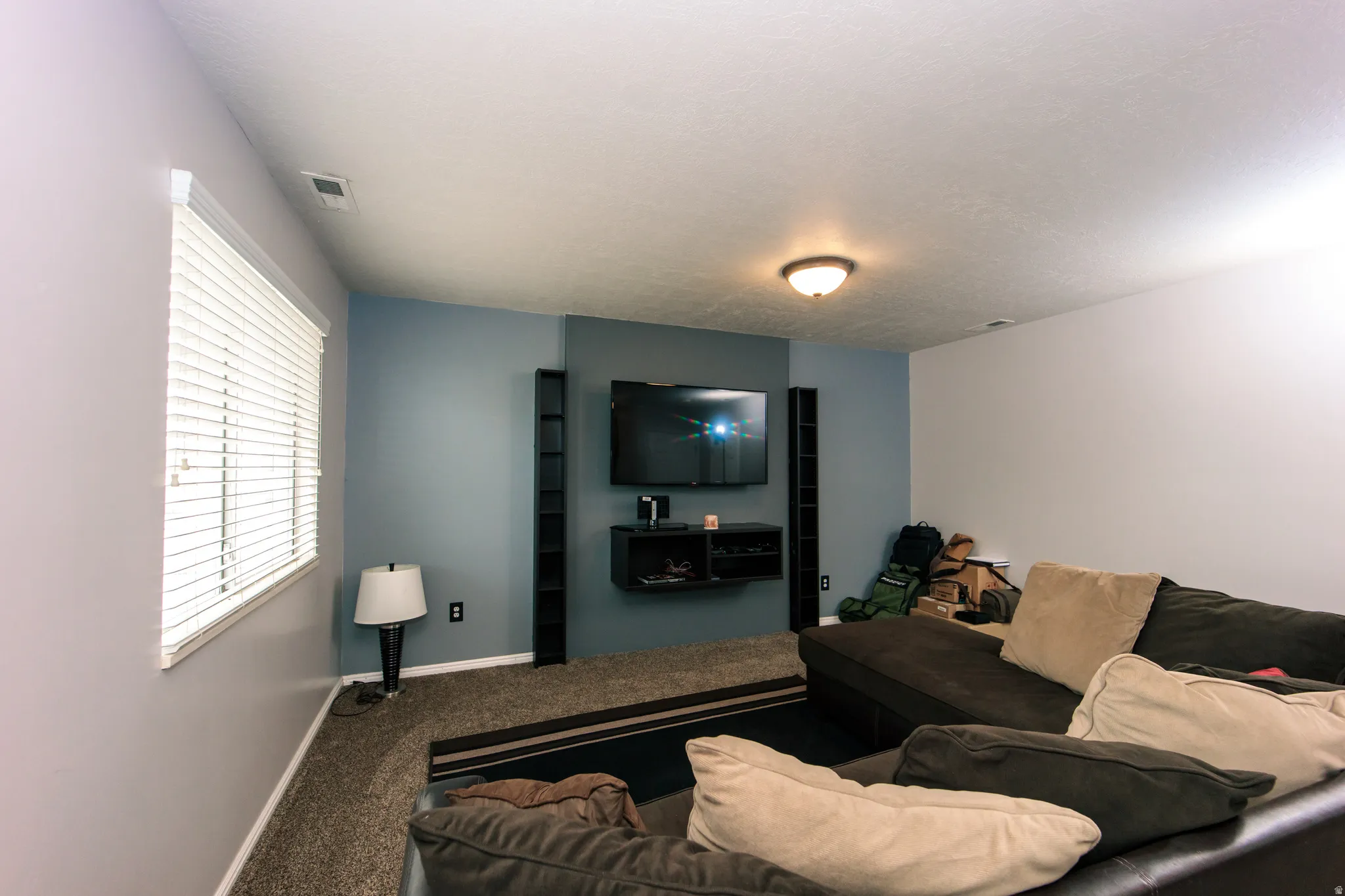Wall-mounted media console, recessed ceiling light, window with horizontal blinds, textured carpet, and a flush-mount ceiling fixture