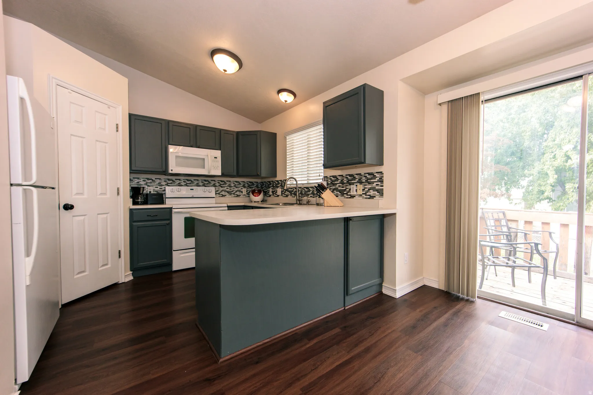 Vaulted ceiling with flush-mount lighting, dark wood-finish flooring, dark cabinetry with white countertops, white appliances, and a sliding glass door to an outdoor deck