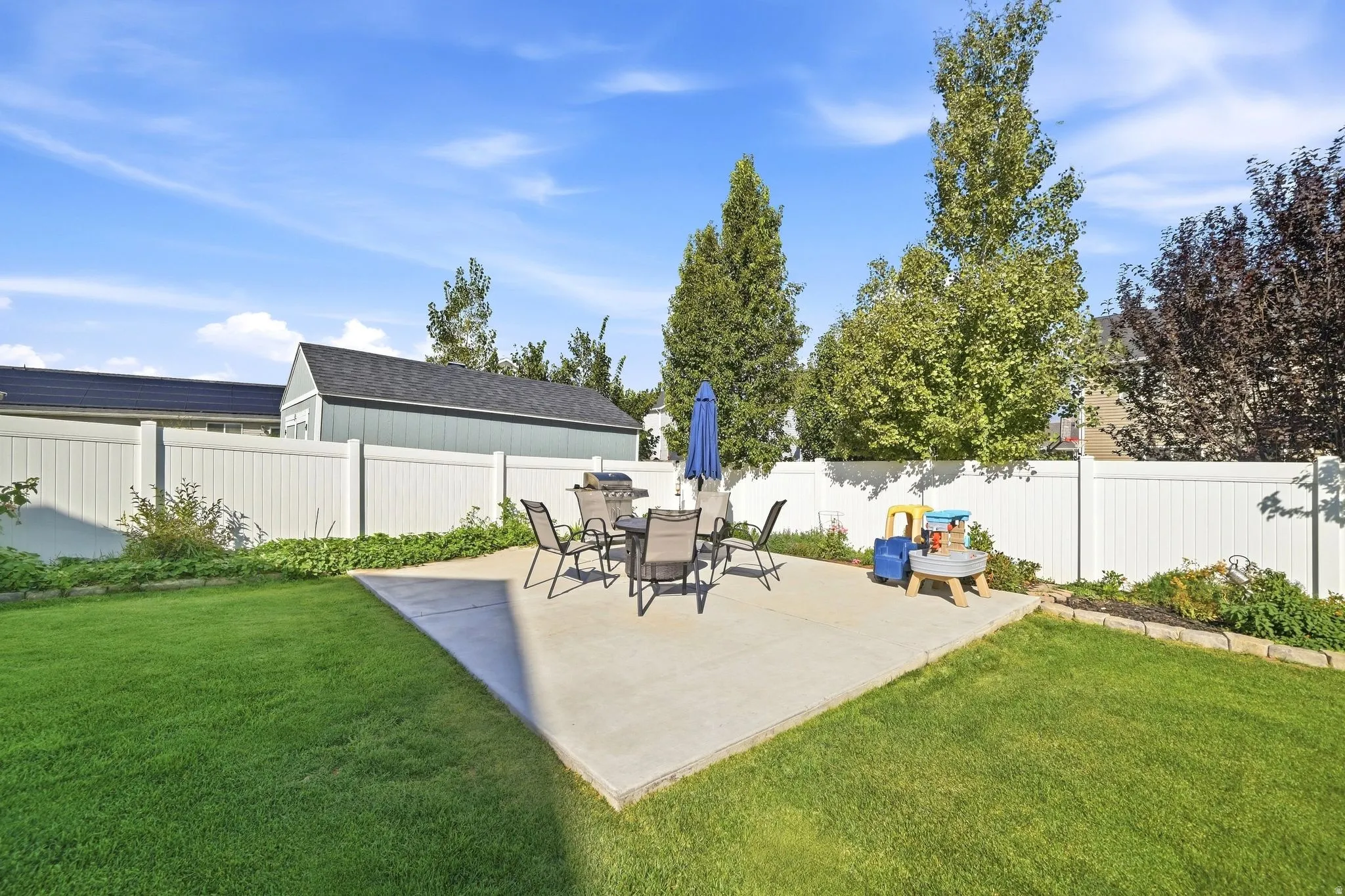 Expansive backyard with a concrete patio, lush lawn, white privacy fencing, and mature trees