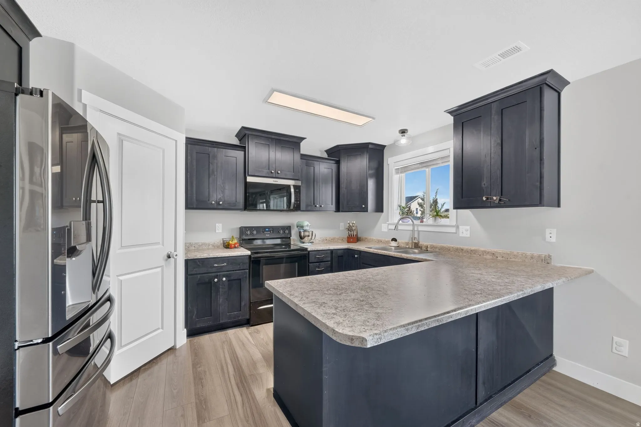 Kitchen featuring wood-finish flooring, dark cabinetry, light-toned countertops, a stainless steel refrigerator, and a window above the sink