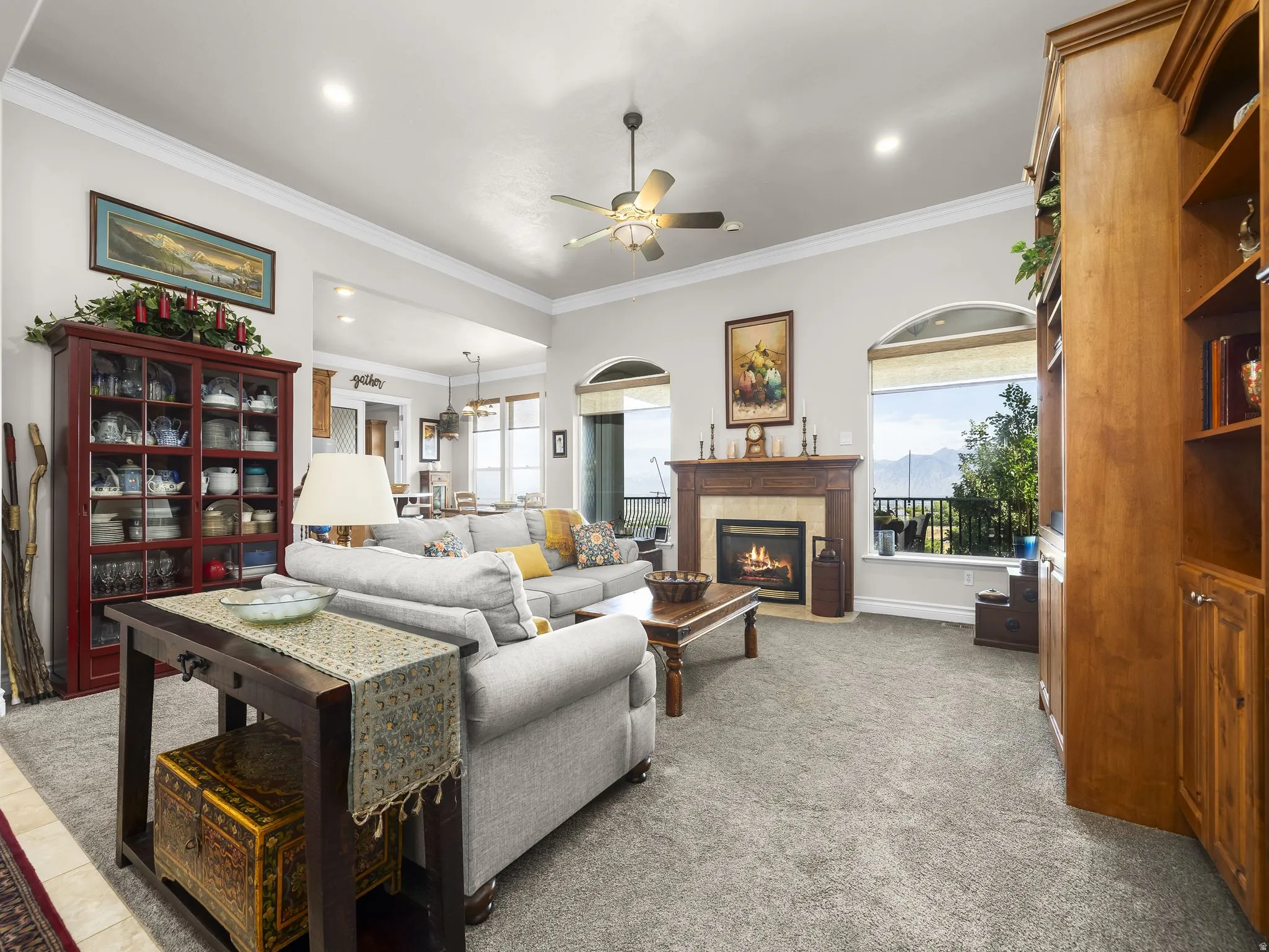 Carpeted living area featuring a fireplace with a wood mantel, crown molding, arched windows, and a ceiling fan