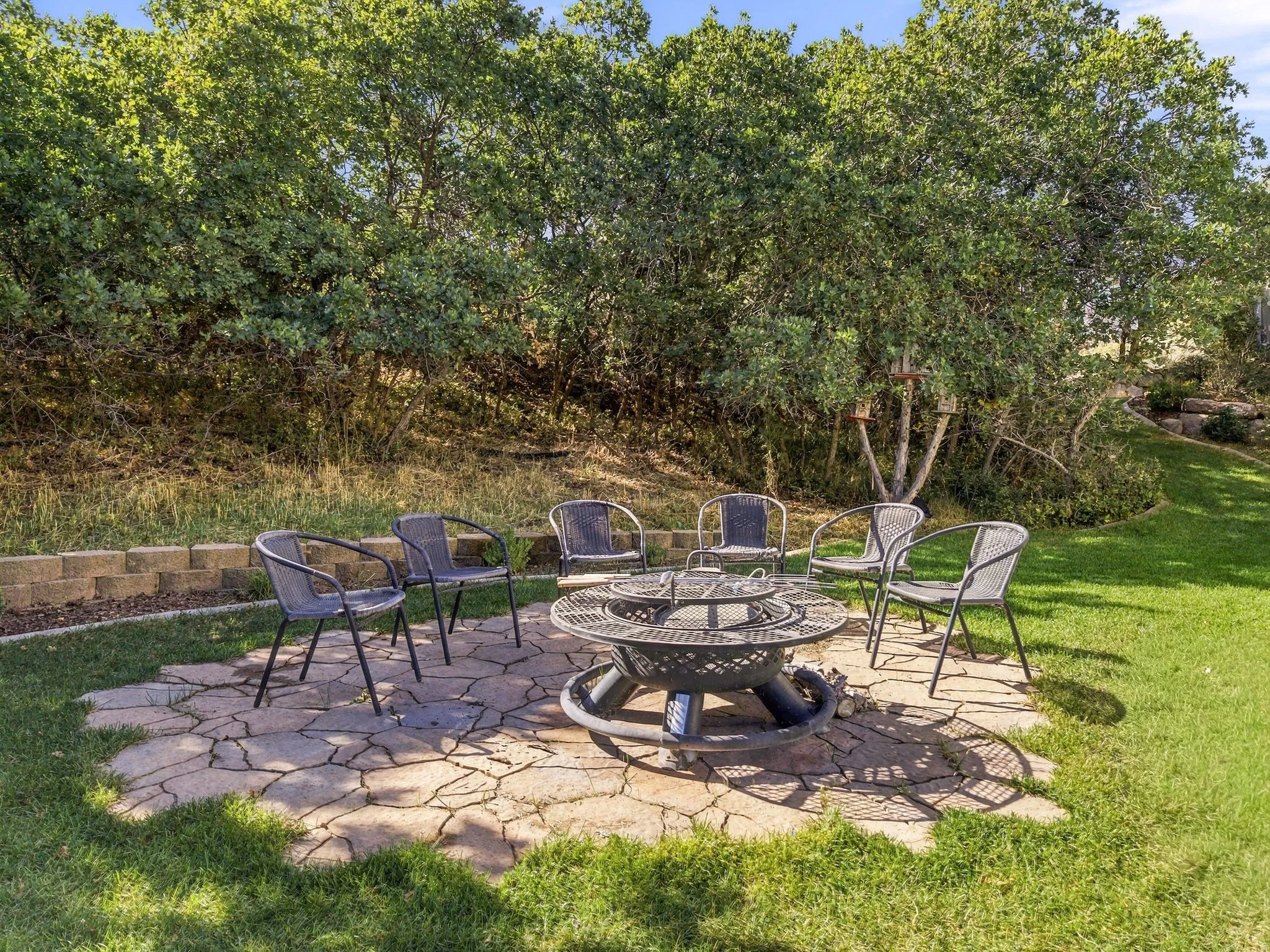 Stone patio with a built-in fire pit, surrounded by a manicured lawn and mature landscaping, and a retaining wall