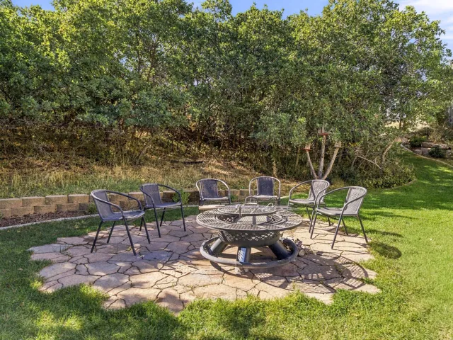 Stone patio with a built-in fire pit, surrounded by a manicured lawn and mature landscaping, and a retaining wall