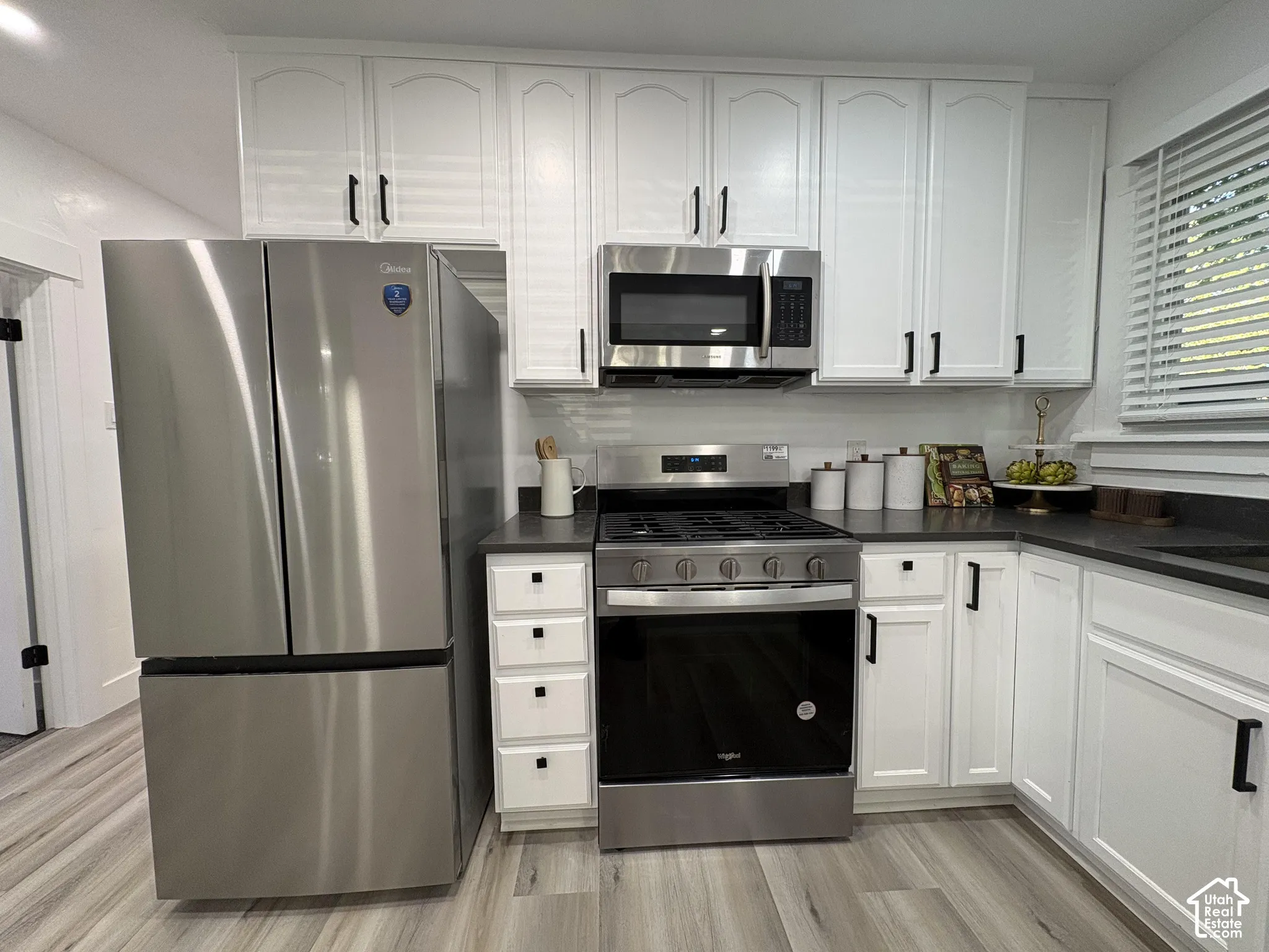 White shaker-style cabinetry with black hardware, stainless steel appliances, dark countertops, wood-finish flooring, and a white tile backsplash