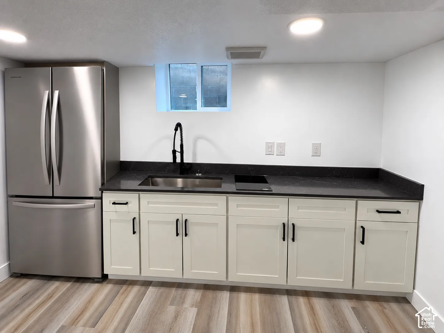 Stainless steel French door refrigerator, dark countertop, single-basin sink with matte black faucet, light-colored cabinetry with matte black hardware, and wood-finish flooring