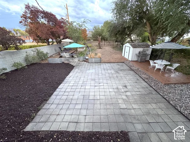 Expansive paved patio with a separate wood deck, raised garden beds, and mature trees