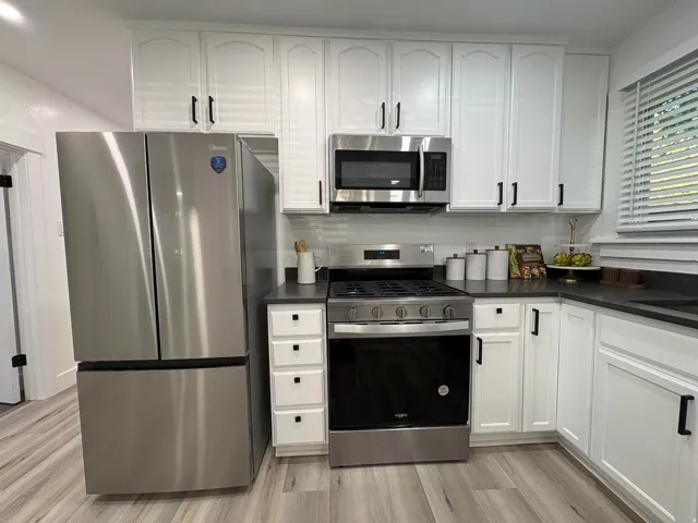 White shaker-style cabinetry with black hardware, stainless steel appliances, dark countertops, wood-finish flooring, and a white tile backsplash