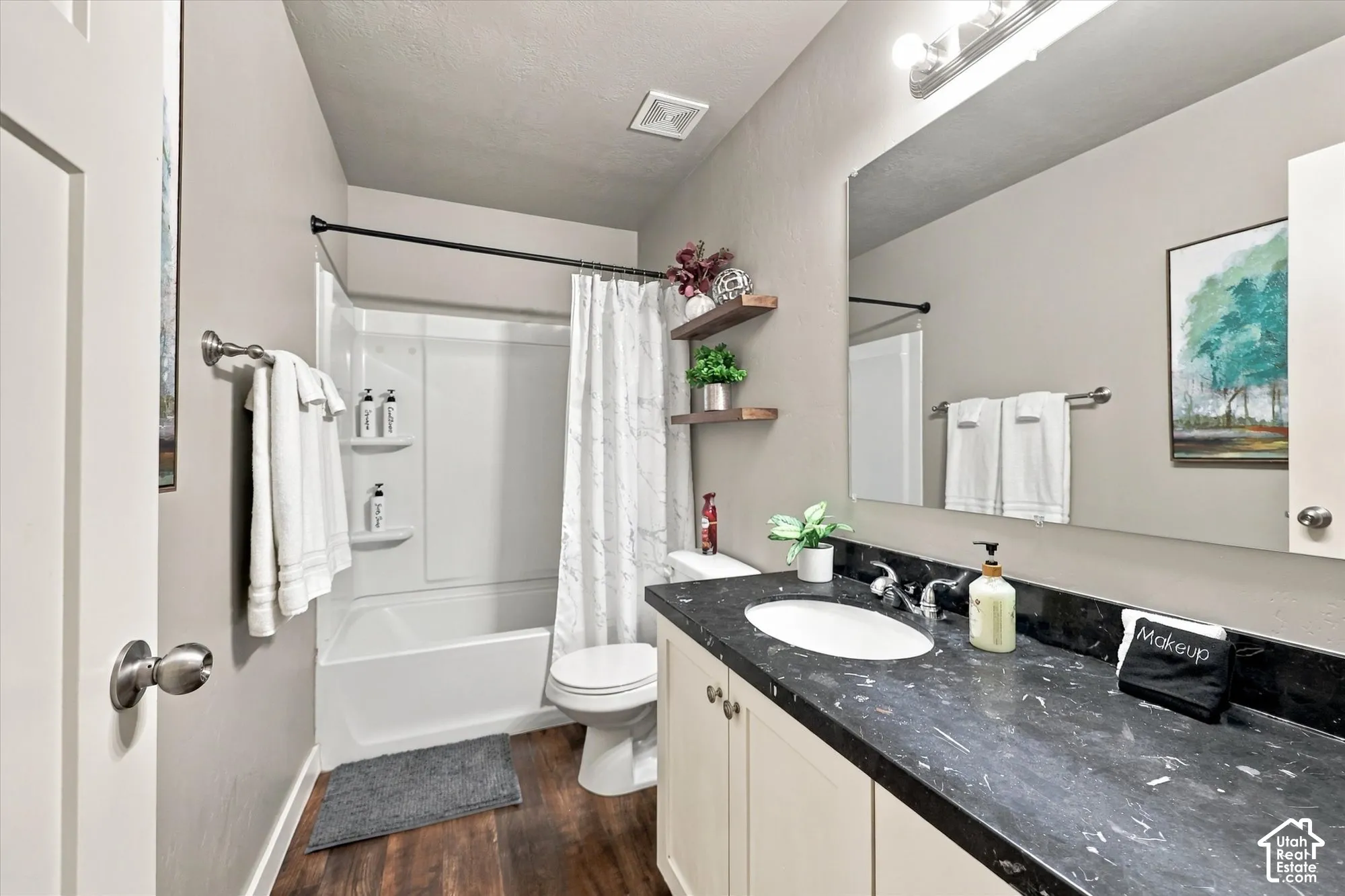 White tub/shower combination, dark wood-finish flooring, white vanity with a dark countertop, single basin sink, and a large wall-mounted mirror
