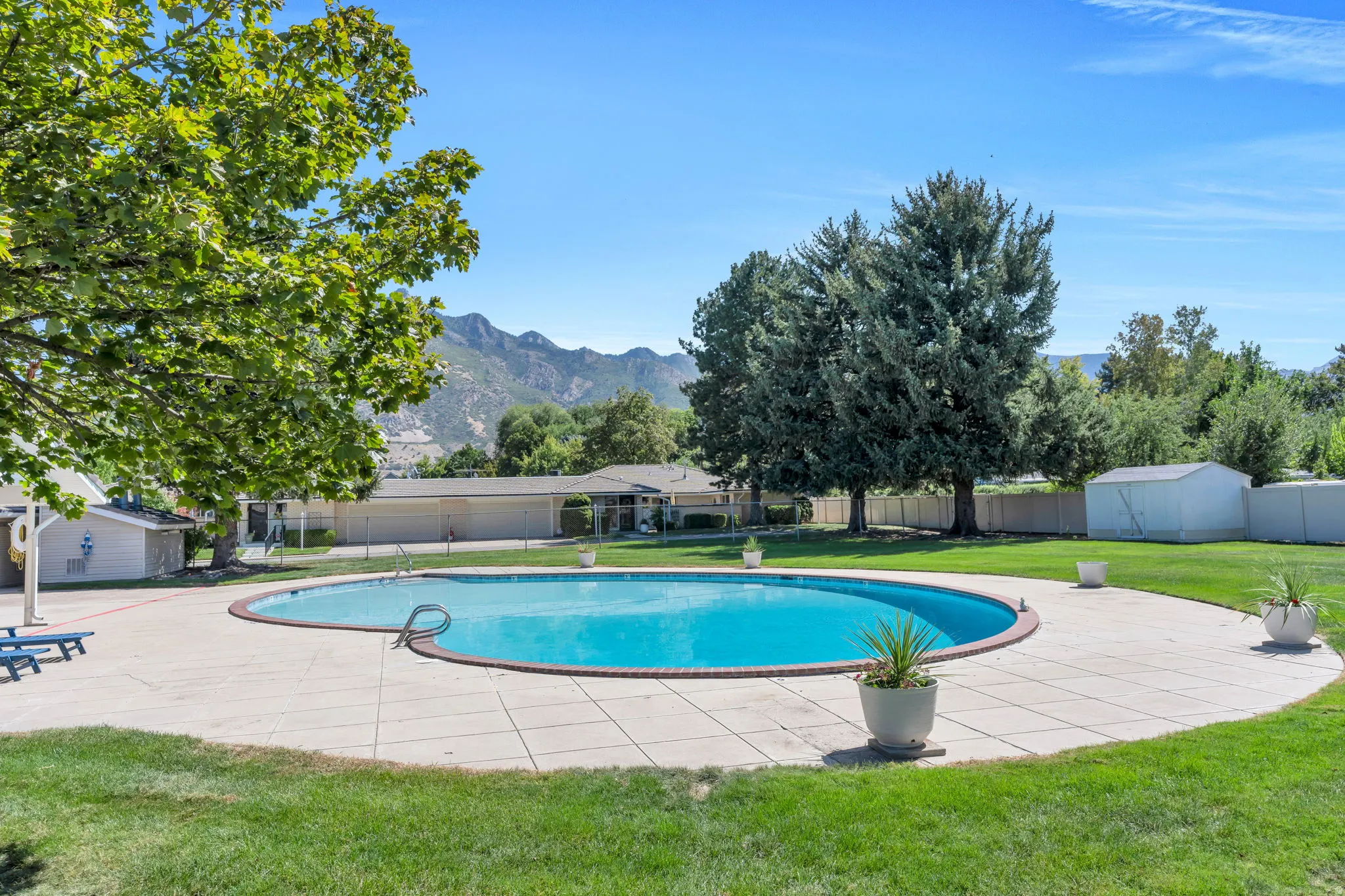 Outdoor swimming pool with surrounding concrete decking, lush green lawn, mature trees, and mountainous background scenery