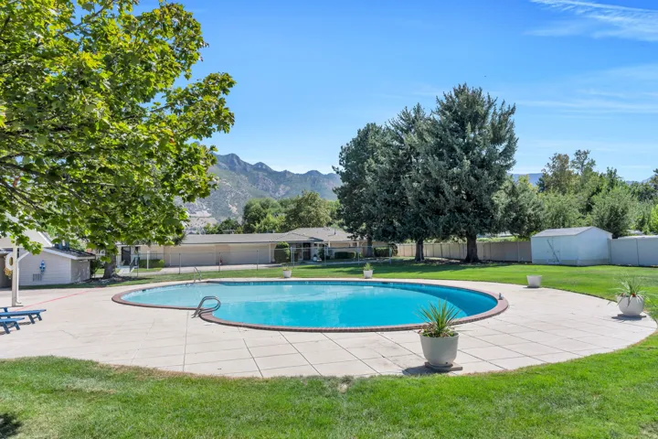 Outdoor swimming pool with surrounding concrete decking, lush green lawn, mature trees, and mountainous background scenery