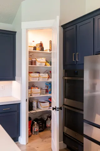 Walk-in pantry with internal lighting, built-in shelving, dark blue cabinetry, light wood-finish flooring, and stainless steel double ovens