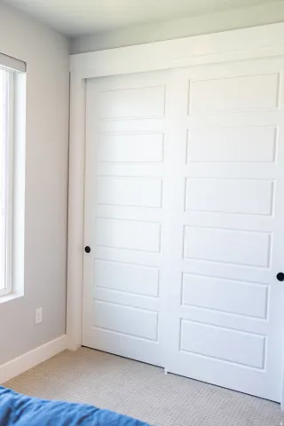 Recessed-panel sliding closet doors with matte black hardware, light-toned carpeting, and a window with a roller shade