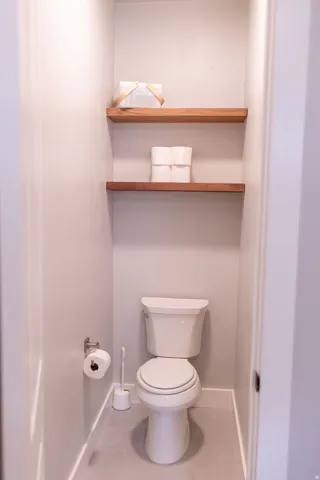 White toilet with a dual-flush mechanism, light gray wall paint, and two wood-finish floating shelves