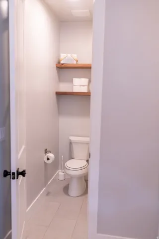 Powder room featuring a white toilet, light gray plank-style floor tiling, and wood-finish floating shelves