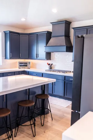 Kitchen featuring dark blue cabinetry, light wood-finish flooring, white countertop surfaces, a built-in range hood, and a tile backsplash