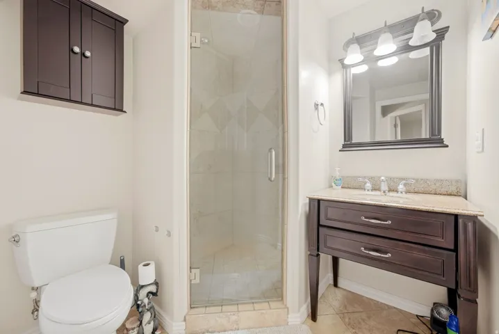 Glass-enclosed shower, dark wood-finish vanity with drawers, granite countertop, wall-mounted mirror, and overhead tri-light fixture