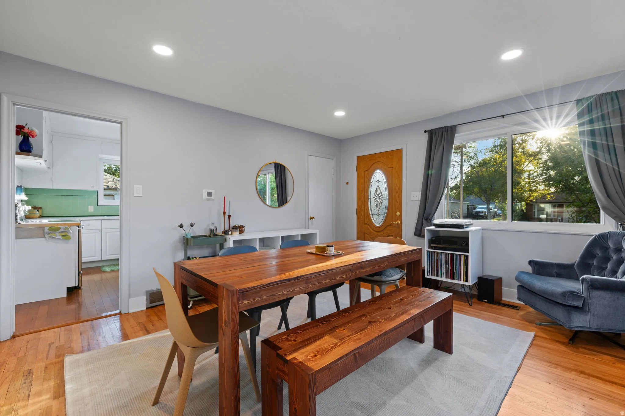Wood-finish flooring throughout, recessed ceiling lighting, a wood-panel entry door with an oval leaded glass insert, and a kitchen with white cabinetry and a green tile backsplash