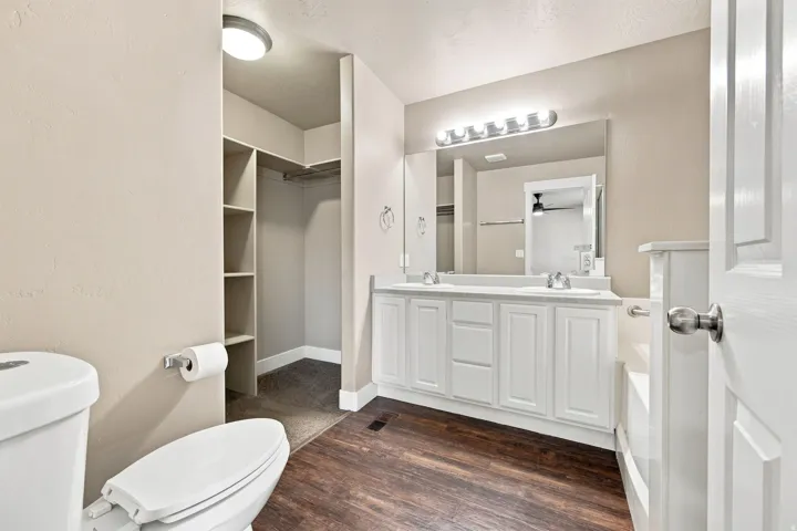 Primary. Wood-finish flooring with a dual vanity, white cabinetry, and an integrated walk-in closet featuring shelving and a hanging rod