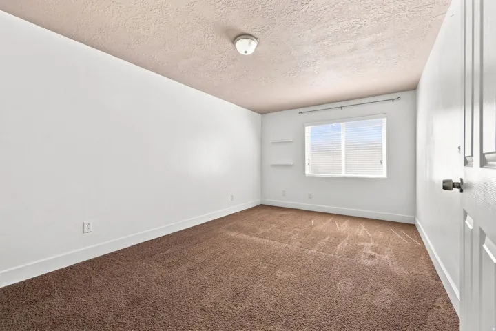 Bed 3. Brown carpeted flooring, white painted walls, a flush-mount ceiling light, a window with blinds, and built-in shelving