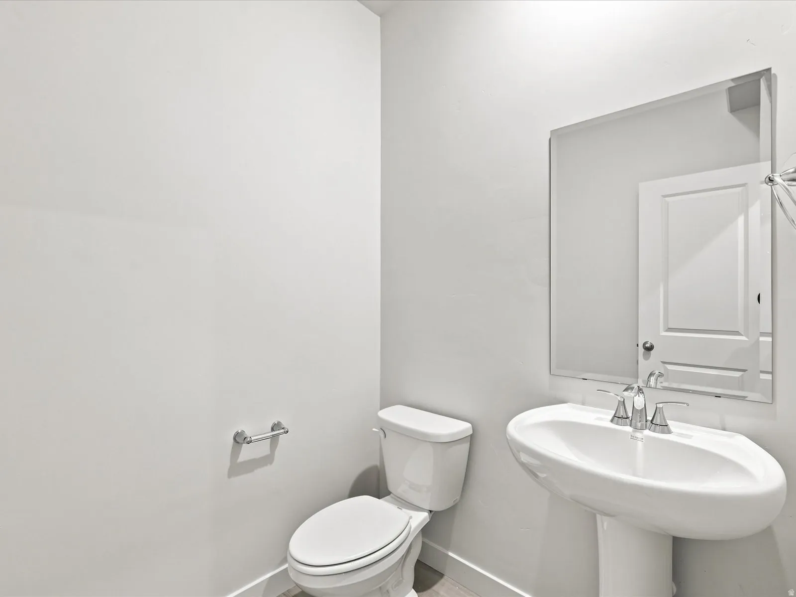 Pedestal sink with chrome fixtures, elongated toilet, large rectangular mirror, and light wood-finish flooring
