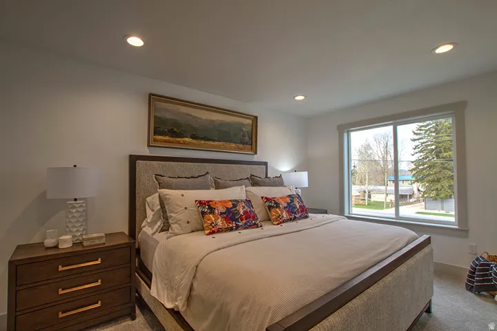 Recessed ceiling lighting, light-toned wall finishes, wide window with white trim, gray carpet flooring, and upholstered bed frame