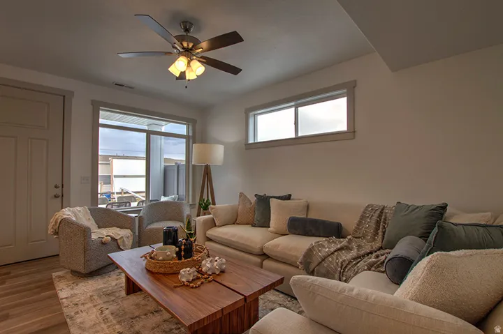 Spacious living area featuring wood-finish flooring, a ceiling fan with integrated lighting, and multiple windows providing natural light