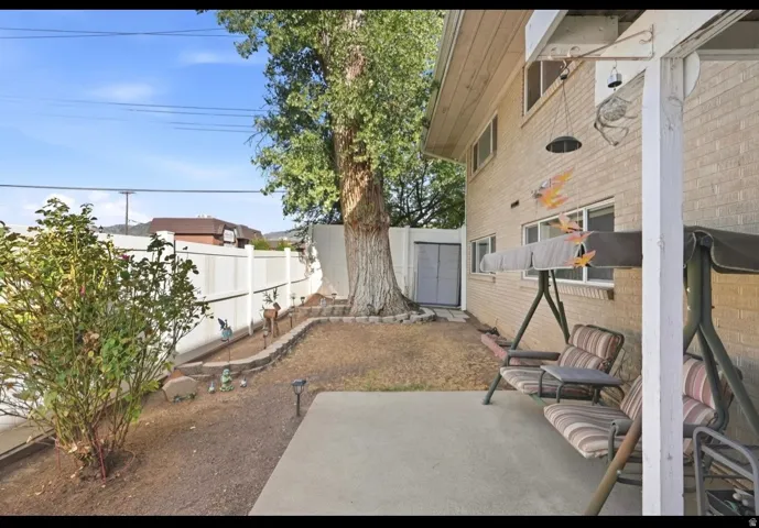 Paved patio area, mature tree, brick exterior, white privacy fencing, and a built-in storage shed