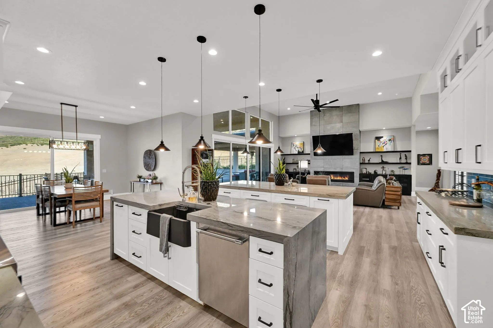 Open-concept living space with wood-finish flooring, white cabinetry, a stone fireplace, and multiple pendant light fixtures