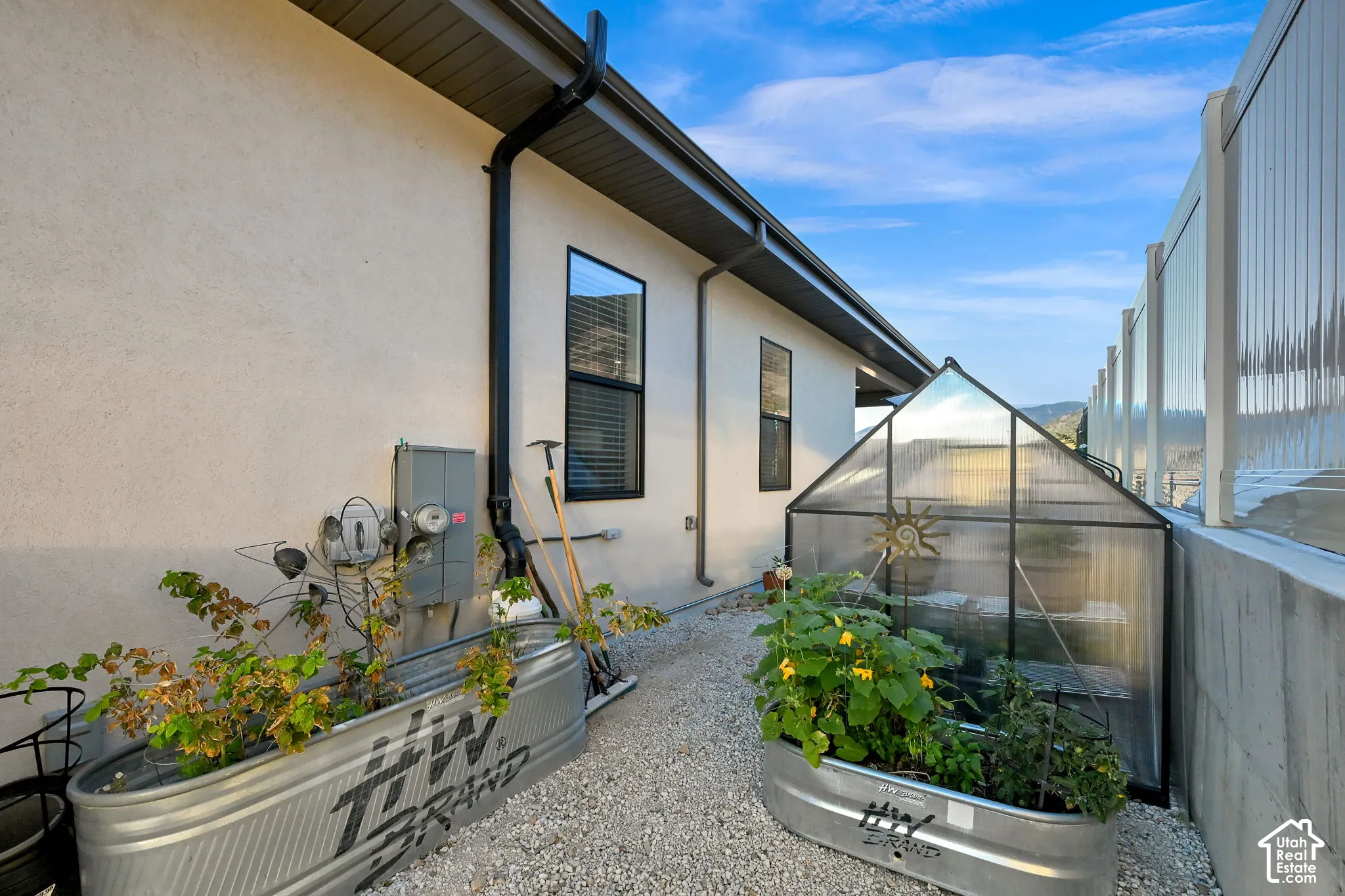 Stucco exterior with black-framed windows, gravel ground cover, and metal raised garden beds and green house.