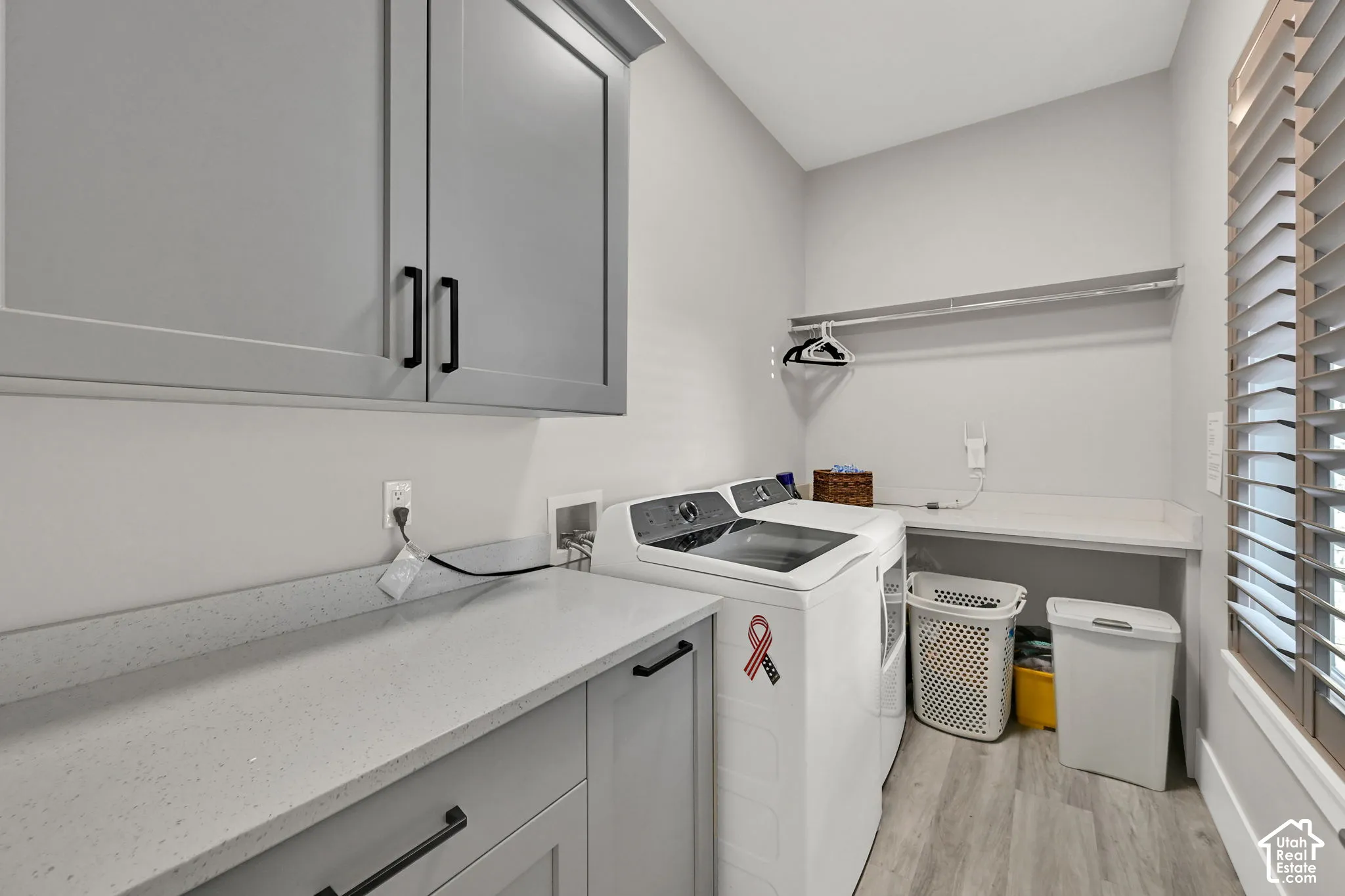 Light gray cabinetry with black hardware, light gray countertops, wood-finish flooring, and a built-in window with Plantation shutters.