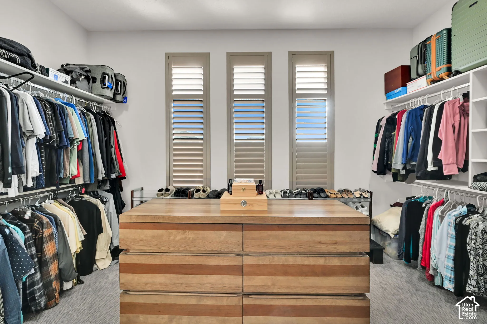 Spacious master walk-in closet featuring central wood-finish island with drawers, extensive wire shelving, multiple hanging rods, and three windows with plantation shutters