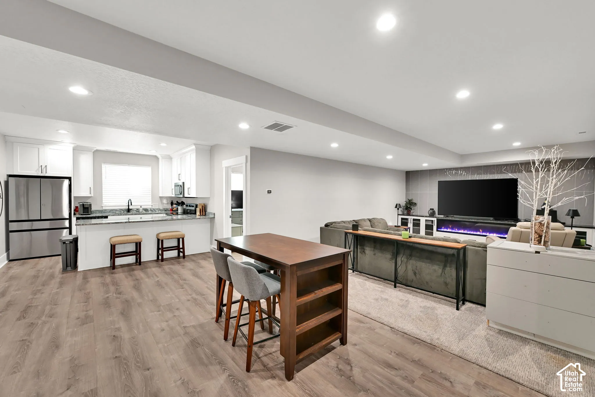Open-concept living space with wood-finish flooring, recessed lighting, white cabinetry, a kitchen island with seating, and a feature wall with a built-in fireplace