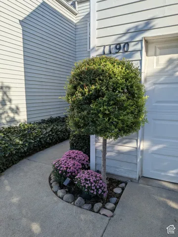 Siding exterior with a clean garage door, concrete walkway, and landscaped planting area featuring a topiary and flowering plants