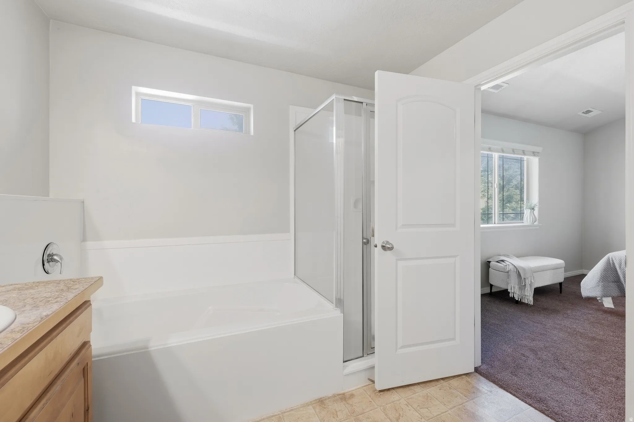 White bathroom featuring a built-in tub, separate glass-enclosed shower, light wood-finish vanity with a countertop, and a window