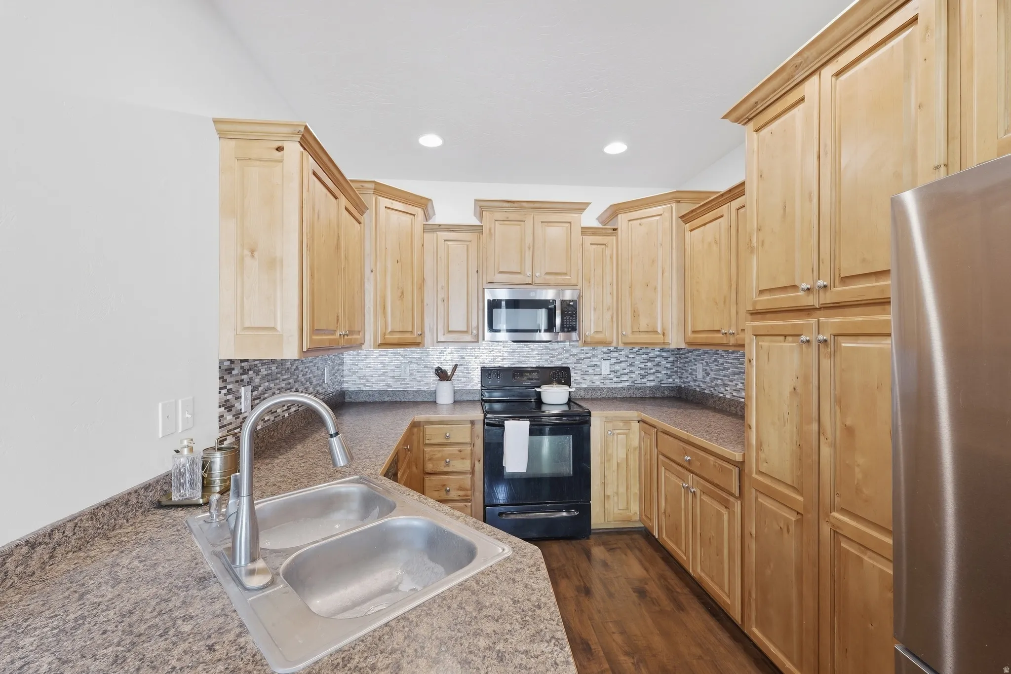 Kitchen featuring light wood cabinetry, stone-finish countertops, stainless steel dual-basin sink with gooseneck faucet, tile backsplash, and wood-finish flooring