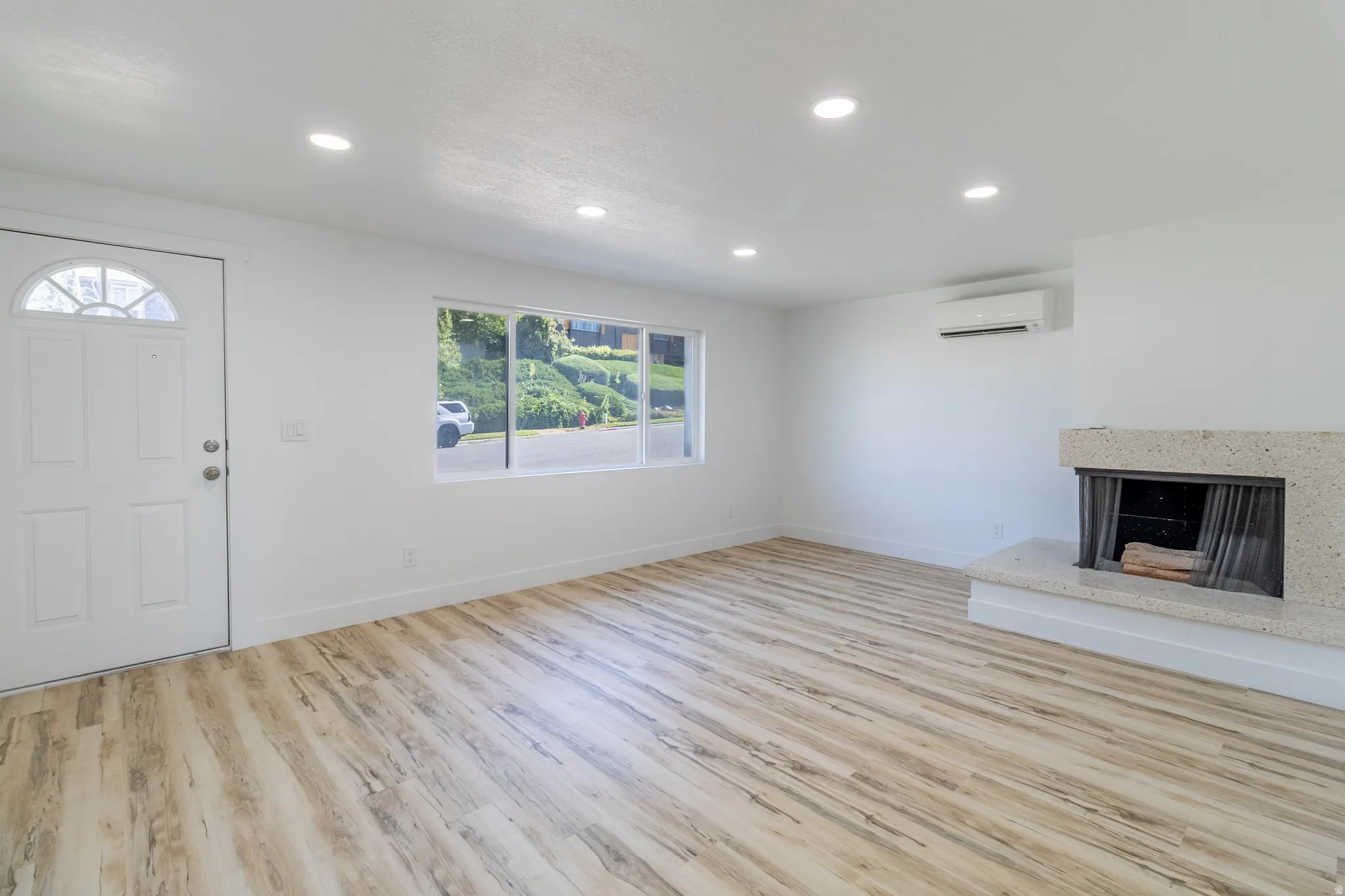 Recessed lighting, light wood-finish flooring, white walls, stone-clad fireplace, and wall-mounted climate control unit