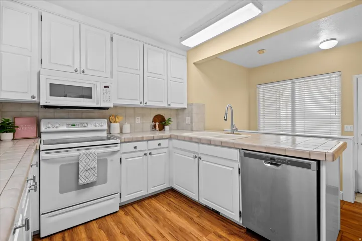White cabinetry with brushed nickel hardware, built-in microwave, stainless steel dishwasher, tile countertops, and wood-finish flooring