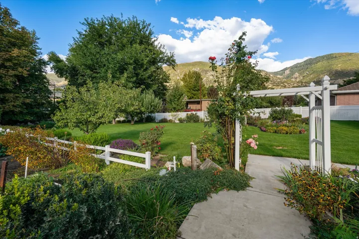 Expansive lawn bordered by mature trees and shrubs, featuring a white fence, garden arbor, and paved walkway