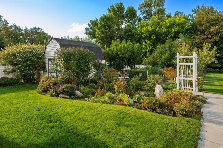 Lush landscaping with diverse plantings, green lawn, garden shed, white picket fence, and a decorative garden arbor