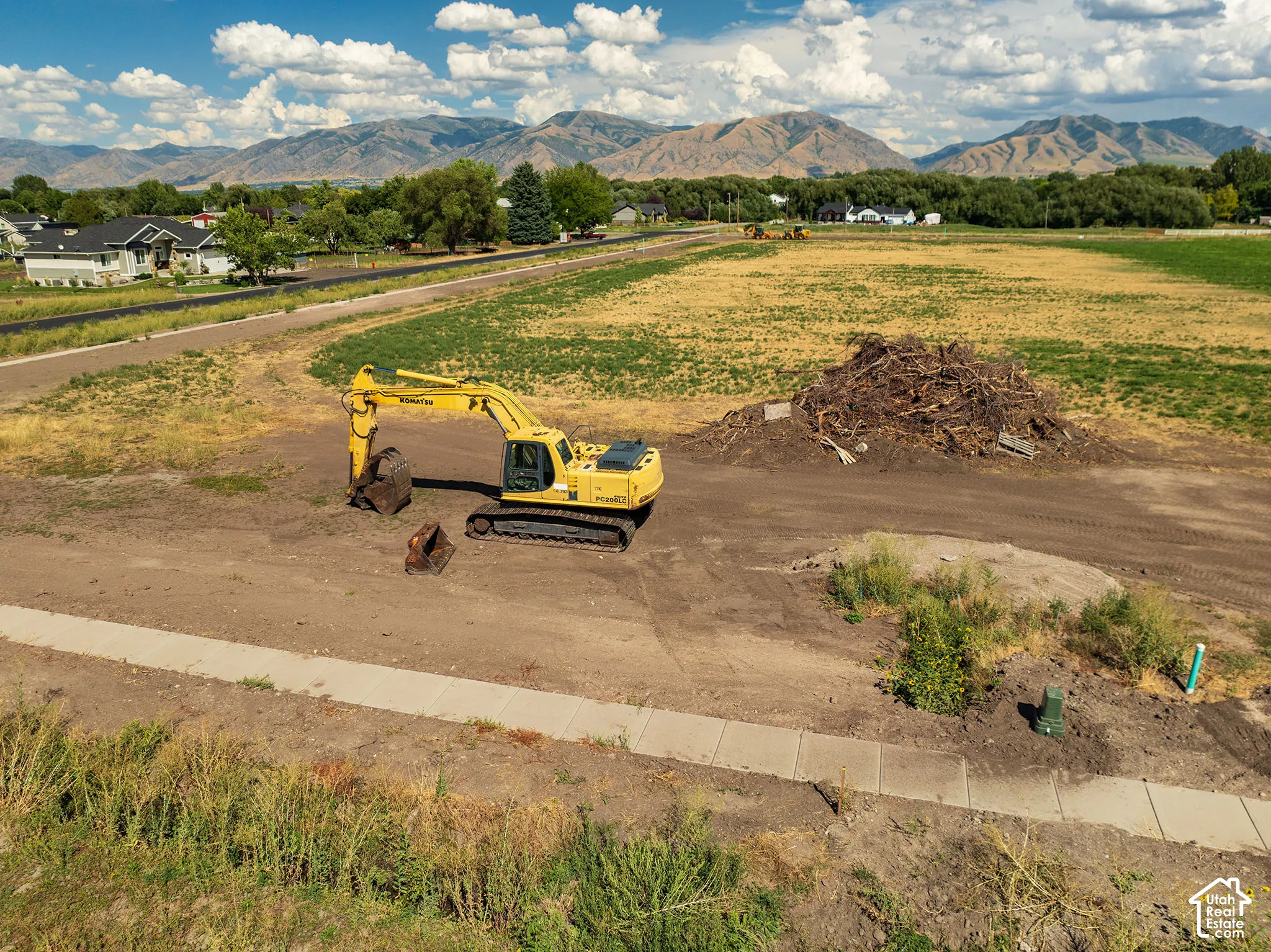 Expansive undeveloped land featuring a paved sidewalk, surrounding residential structures, and distant mountains