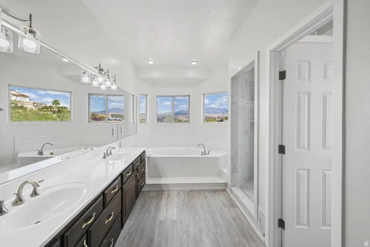 Dual vanity with undermount sinks, wood-finish flooring, a built-in tub, and a glass-enclosed shower with tiled walls