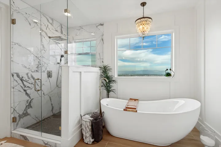 Freestanding soaking tub with a large window, glass-enclosed shower with marble-look tile, and wood-finish flooring