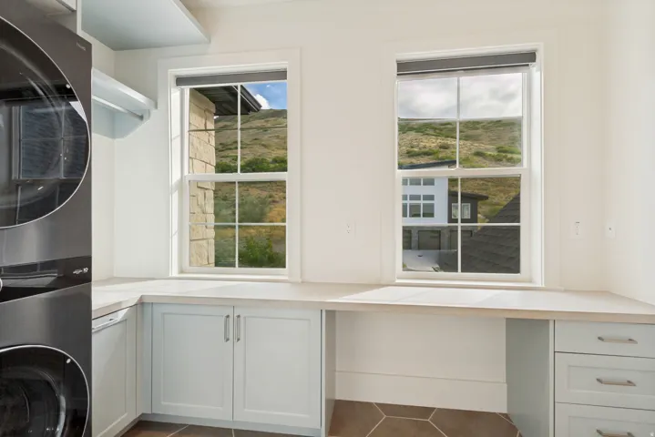 Dual windows with roller shades, light-toned countertops, light blue shaker-style cabinetry, and hexagonal floor tiling