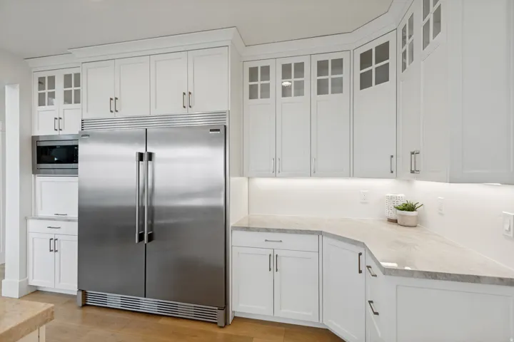White shaker-style cabinetry, stainless steel refrigerator, built-in microwave, under-cabinet lighting, and light-toned countertops