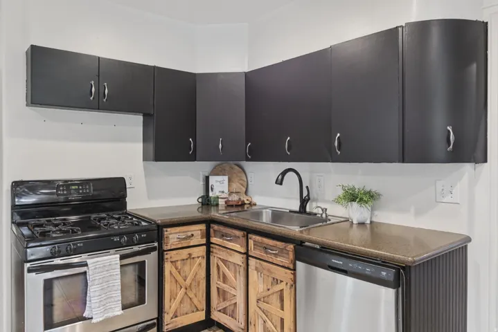 Stainless steel gas range, integrated stainless steel dishwasher, black upper cabinetry, wood-finish lower cabinetry, and a dark countertop